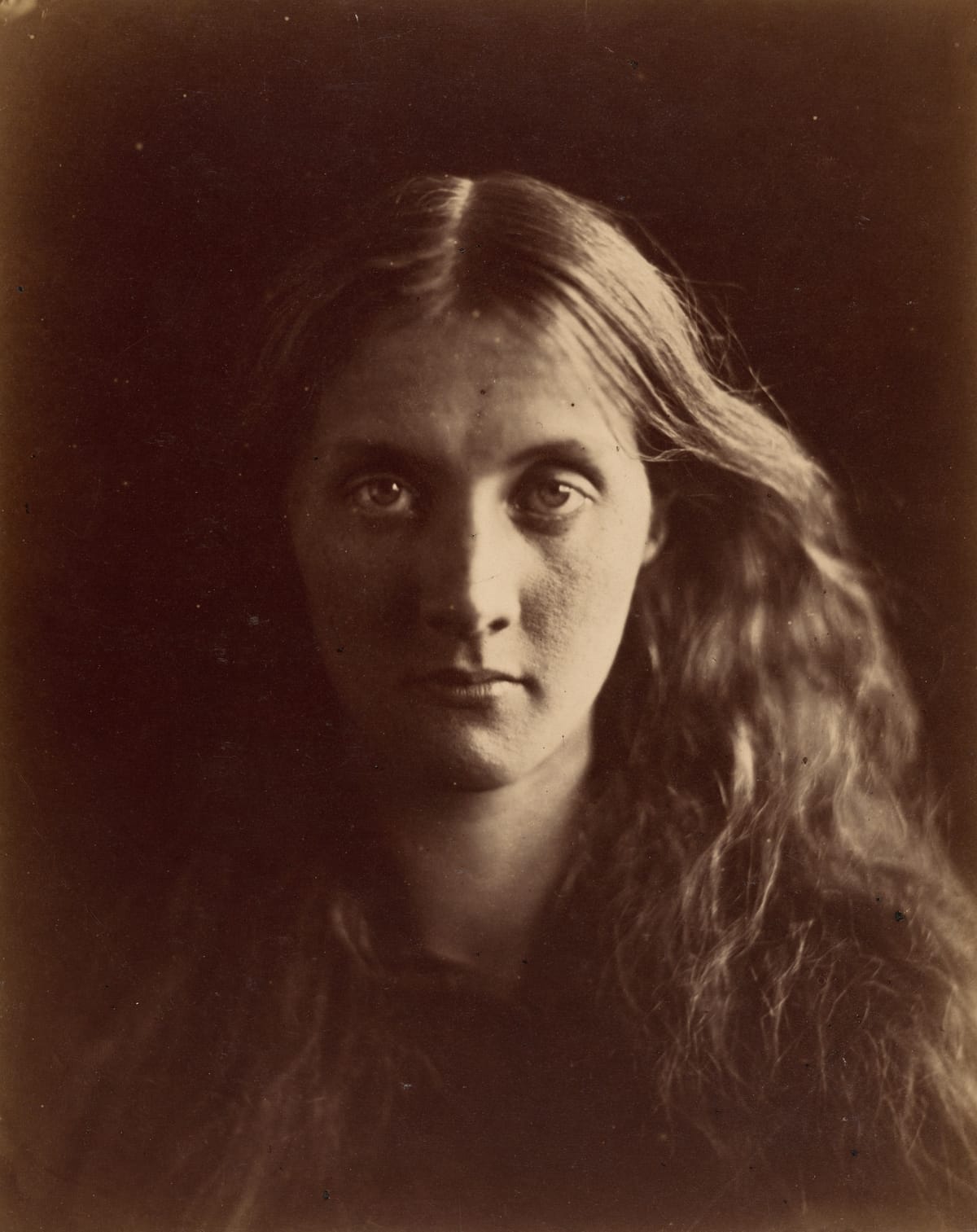 Sepia photograph of a young woman with long, loose, wavy hair parted in the middle, looking straight at the camera, her face half lit against a dark background.