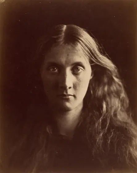 Sepia photograph of a young woman with long, loose, wavy hair parted in the middle, looking straight at the camera, her face half lit against a dark background.