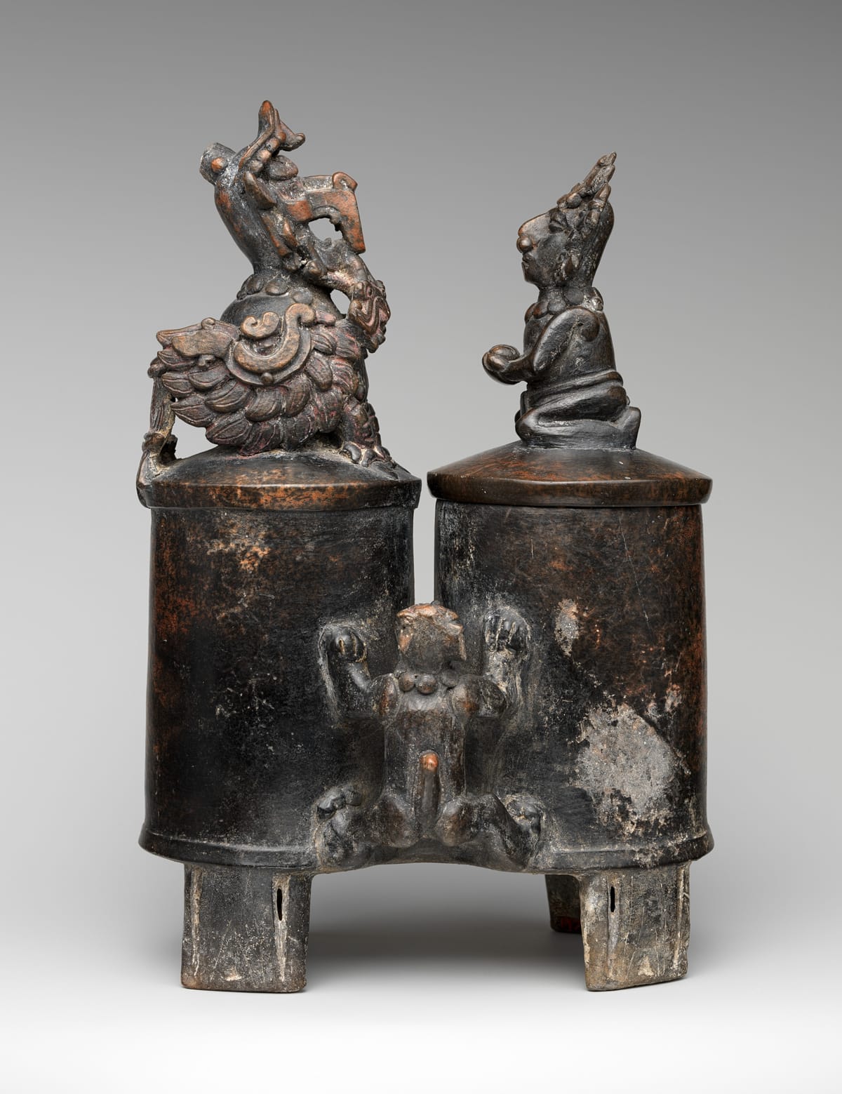 Dark burnished ceramic vessel of two joined cylinders standing on slab feet; each lid is topped by a small sculpted figure, and a modelled figure clings to the join between the two chambers.