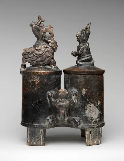 Dark burnished ceramic vessel of two joined cylinders standing on slab feet; each lid is topped by a small sculpted figure, and a modelled figure clings to the join between the two chambers.