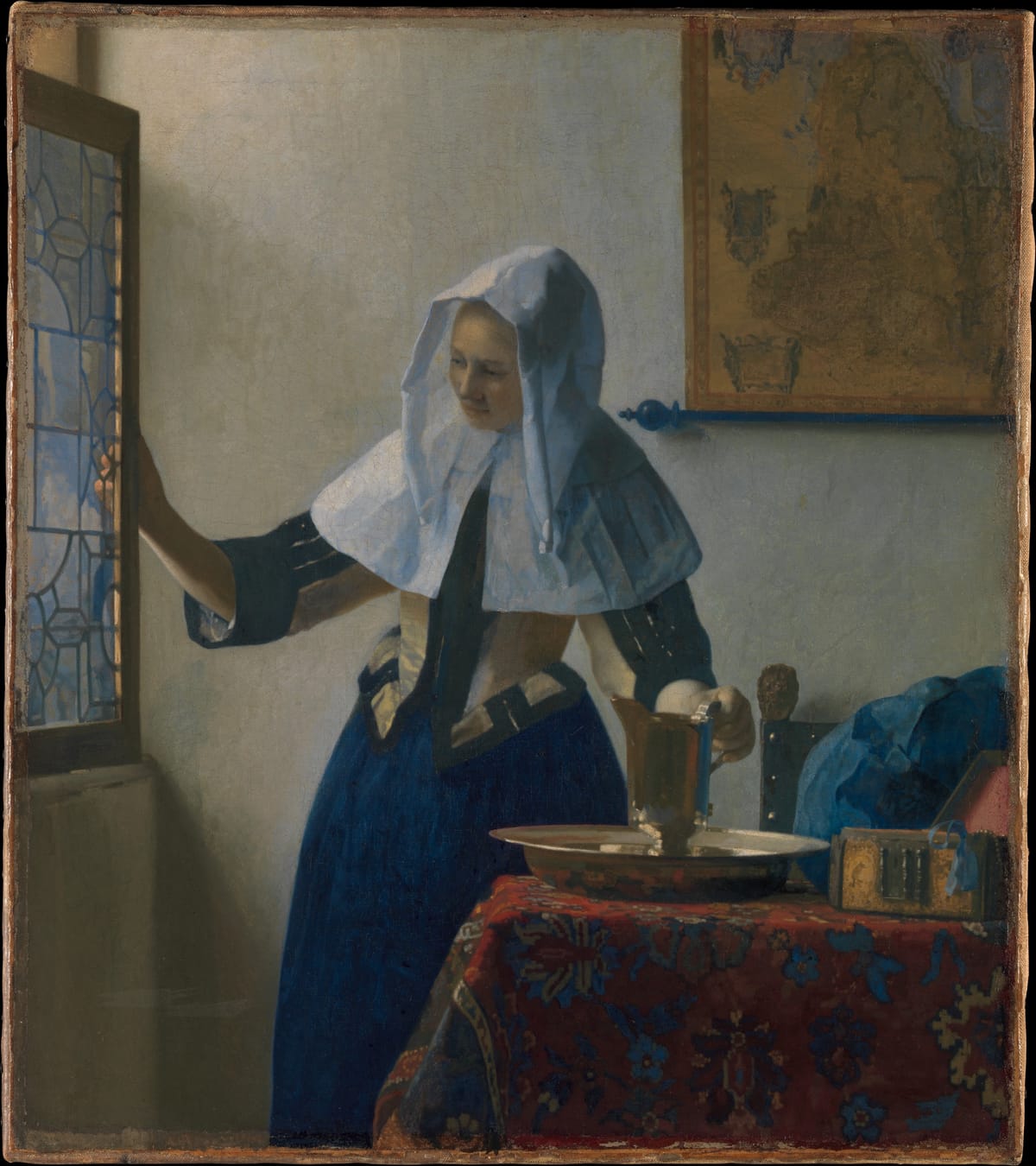 A young woman in a white linen hood and collar, yellow bodice and blue skirt stands at a leaded-glass window, one hand opening the casement and the other resting on a gilt silver pitcher in a matching basin on a table covered with a red patterned carpet; a map hangs on the pale wall behind her.