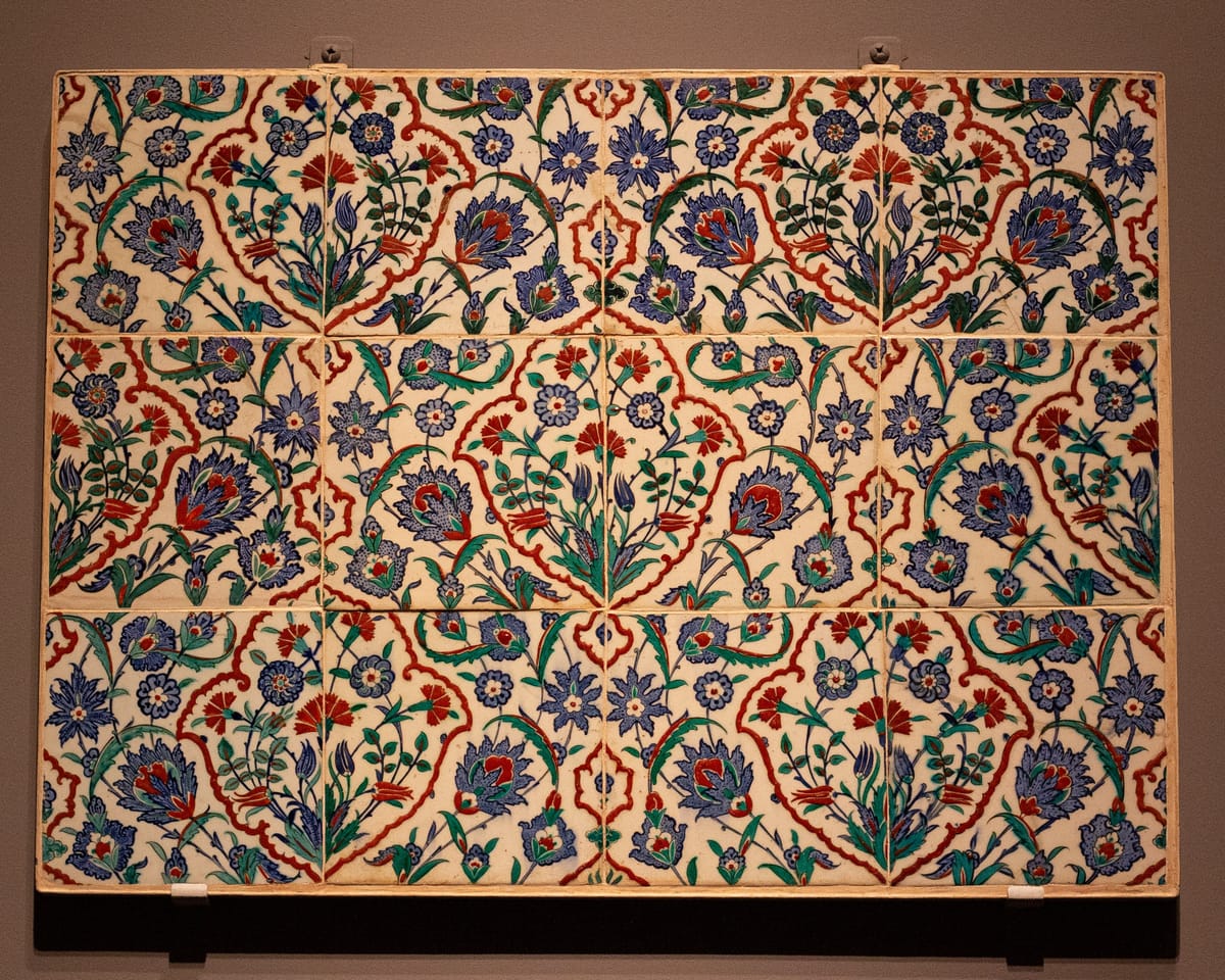 Panel of glazed tiles with a repeating pattern on a white ground: red-outlined medallions and curving leaves frame blue and red flowers, carnations and tulips on green stems.