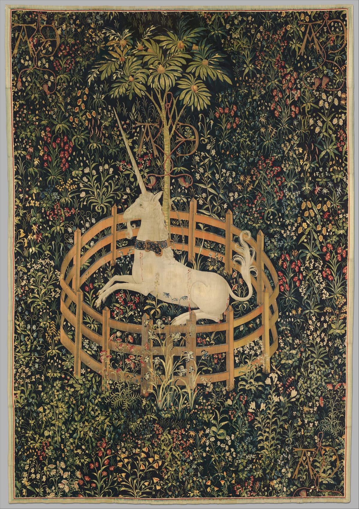 Tapestry of a white unicorn with a gold collar lying inside a low, round wooden fence, chained to a fruit-laden pomegranate tree, against a dense dark-green ground filled with flowers and plants.